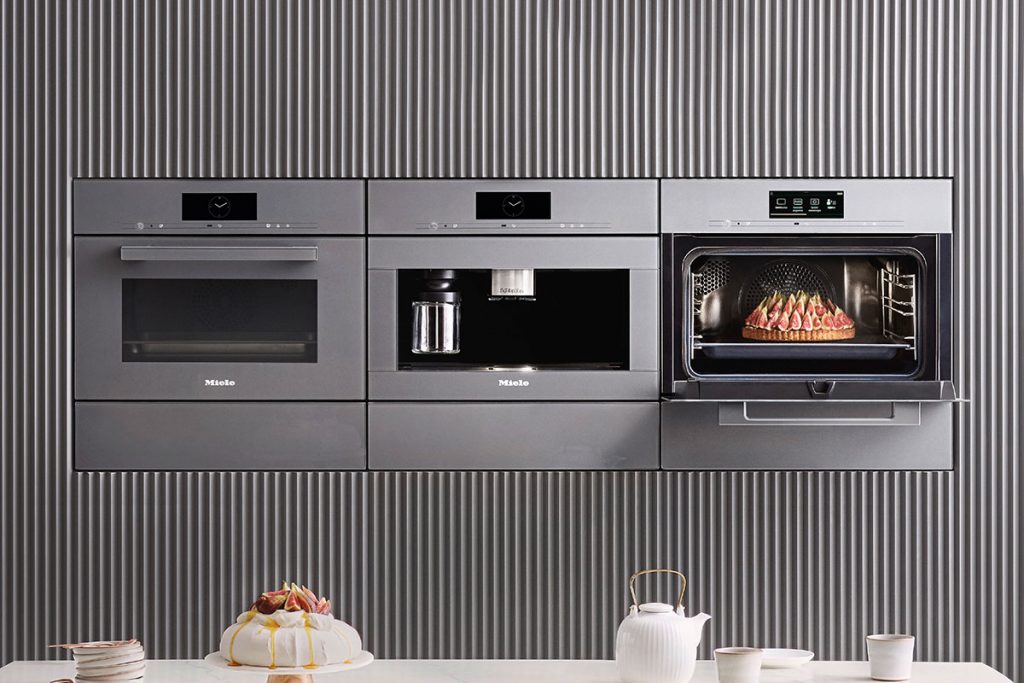 a row of three stainless steel Miele ovens, two closed and one with a Pavlova dessert on display, in front of an island set for a party, to illustrate a feature on kitchen must-haves for entertaining