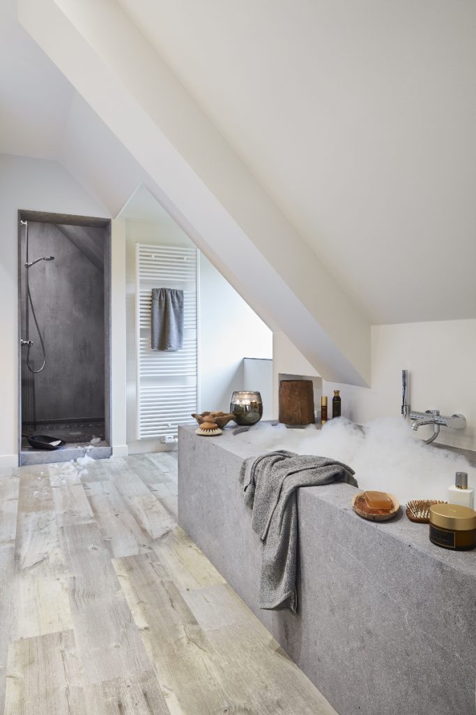 a modern rustic bathroom featuring a grey stone bath filled with bubbles, with accessories around it including candles and a soap dish